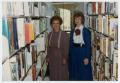 Photograph: [People Standing Between Library Bookshelves]