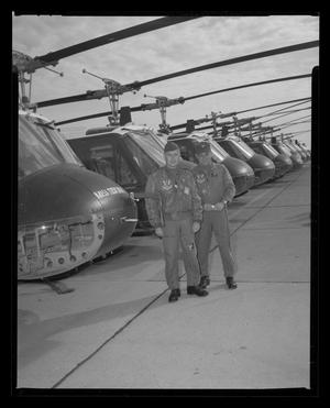 Primary view of [Photograph of two men posing in front of a row of UH-1F Iroquois helicopters]